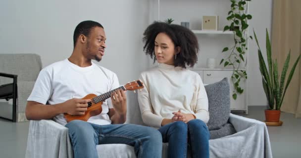 Afro american couple newlyweds sitting on sofa in cozy living room black man holding musical instrument ukulele guitar talking with beloved wife discussing smiling going to sing song, hobby concept — Stock Video