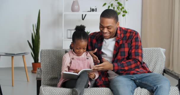 Afro amerikansk far hjälper älskad rolig dotter att läsa grå bok uppmanar liten flicka arg täcker hennes pappor mun med handen kräver tystnad, etnisk familj skrattar i vardagsrummet, hem utbildning — Stockvideo