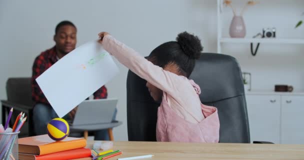 Little cute Afro American daughter school girl draws picture with colored pencils while her father typing on laptop at home during quarantine baby shows paper to her dad man stops work supports child — Stock Video