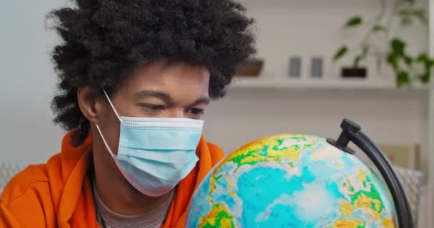 Retrato de afroamericano con máscara médica mirando el mapa del mundo del globo despega respirador sonriente elige país donde ir a viajar después de bloquear cuarentena cancelada, fin de pandemia — Vídeo de stock