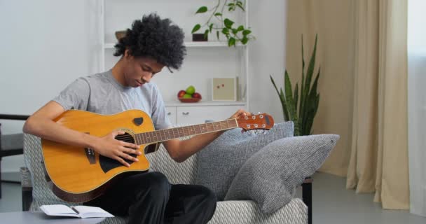 Afro-americano menino adolescente músico homem negro artista sentado no sofá em casa afinando tom de corda de guitarra soando se preparando para tocar instrumento musical lição de prática musical em casa, passatempo criativo — Vídeo de Stock