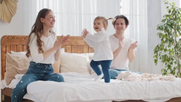 Gelukkig gezin jonge ouders met baby kleine dochter zuigeling zitten op bed, klein meisje peuter kind dansen op muziek moeder en vader applaus klappen handen ondersteuning kind dans, leuke vrije tijd in huis slaapkamer — Stockvideo