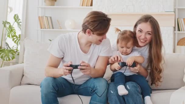 若い家族は家のソファ隔離レジャーで座っているオンラインビデオゲームをプレイします,思いやりのある楽しい両親のお母さんとお父さんcucasianお母さんとお父さんは小さな赤ちゃんの小さな娘かわいい幼児の遊びコンソール — ストック動画