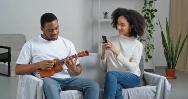 Seniman musisi Afroamerika memainkan gitar kecil bernyanyi melihat kamera ponsel gadis keriting menggunakan perangkat smartphone untuk merekam video untuk jejaring sosial mengambil foto musisi favoritnya — Stok Video