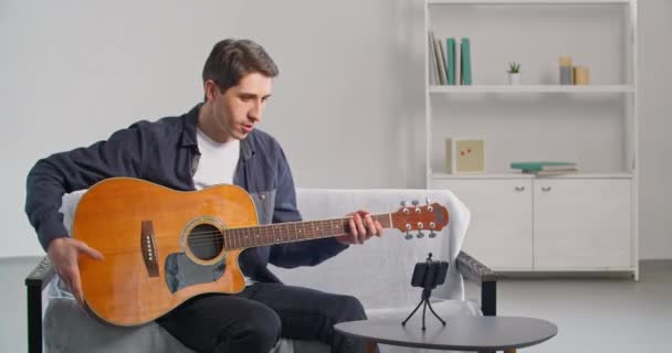 Vit kille sitter på soffan använder stativ och smartphone för att spela in video lektion för blogg på nätverk visar gitarr på kameran förklarar spelteknik musikinstrument, modern konst utbildning online — Stockvideo