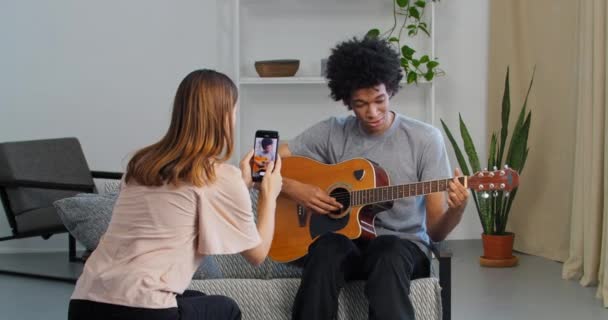 Afro american guy teen blogger playing guitar at home speaks explains teaches remotely online while caucasian woman friend helps record video lesson on mobile phone for social networks during lockdown — Stock Video