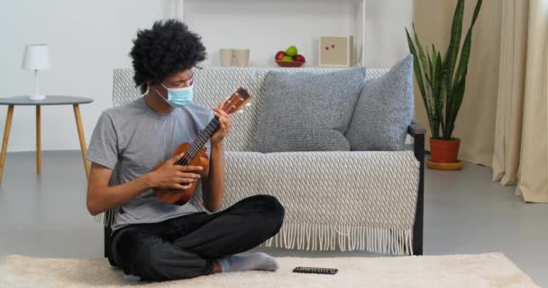 Afro Američan chlap nemocný muž hudebník nosí ležérní oblečení a lékařskou masku obličeje sedí na podlaze hraje ukulele malé kytara hudební nástroj praktikující hudbu doma během karantény a uzamčení — Stock video