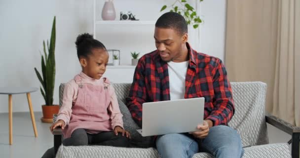 Young father afro american guy sits on couch with little cute girl his daughter teaches child how to use laptop play video games going to watch movie or shopping with baby calling her to move closer — Stock Video