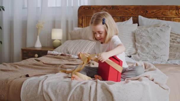 Curious little girl happy preschooler blonde daughter cute baby kid child sitting on cozy bed with festive red gift box unpacks present unties yellow ribbon looks inside, home holiday birthday party — Stock Video