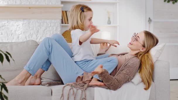 Söt söt liten barn dotter sitter på ung mamma liggande på soffan och leker talande hålla händer hemma, lycklig familj mor och små barn förskola flicka njuter binda ha kul på soffan — Stockvideo