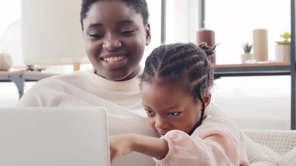 Portrait femme mère africaine et afro américaine petite fille noire enfant assis ensemble à la maison regardant dans un ordinateur portable, enfant pointe du doigt à l'écran saluant, famille faisant appel vidéo en ligne — Video