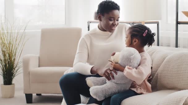 Mãe africana feliz com afro pequena filha estudante sentada no sofá em casa conversando conversa, mulher negra mãe babá se comunicando conversando com criança menina pequena segurando ursinho de pelúcia — Vídeo de Stock