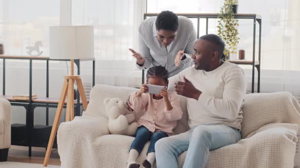 Afro amerykański mężczyzna ojciec dziadek z małą córeczką dziecko gra w mobilne gry wideo online przez telefon siedzi na kanapie młoda czarna matka wściekła Afrykanka kobieta krzyczy przysięga, konflikt rodzinny — Wideo stockowe