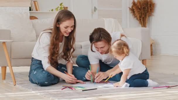 Feliz lazer em casa de família, os pais jovens desenho colorir imagem com lápis deitado no chão ajudando a criança bebê bonito pequena filha desfrutando do jogo, pai mãe e menina criança pequena se divertindo interior — Vídeo de Stock