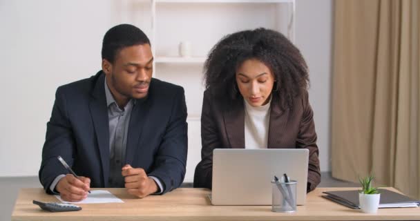 アフリカ系アメリカ人女性の金融コンサルタント会計士は、費用の民族のビジネスマンのコスト会計を助ける会社の計画予算は、ノートパソコンに見えるの数は、文書で報告書を書く話す男を指示 — ストック動画