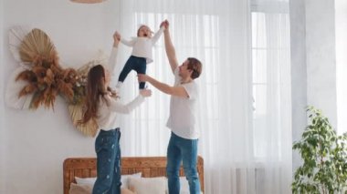 Genç anne ve beyaz anne ve bin yıllık baba evde yatakta duruyor, küçük kız çocuğunu kaldırıyor küçük kız çocuğu elleriyle havadan havaya kaldırıyor aktif olarak hafta sonu birlikte oyun oynuyorlar.