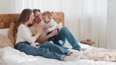 Küçük kızıyla birlikte beyaz bir anne ve küçük bir bebek rahat bir yatakta uzanıyorlar, anne bebekle pasta oyunu oynuyorlar kahkahalarla gülüyorlar, mutlu bir aile ve çocuk yatak odasında dinleniyorlar.
