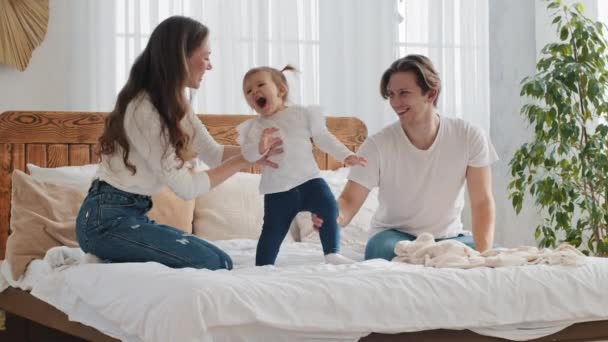 Happy family young parents with baby small daughter have fun playing jumping on bed cozy bedroom, caring mother holds active child little infant toddler, kid yells from fatigue, father and mom laugh — Stock Video
