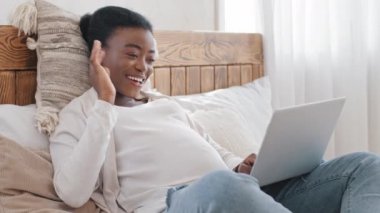 Afrikalı mutlu, hamile, serbest çalışan, bilgisayarlı, gelecekteki yalnız anne afro kız. İnternetten sohbet görüşmesi yapıyor. Uzaktan evden yatağa uzanmış, el sallıyor.