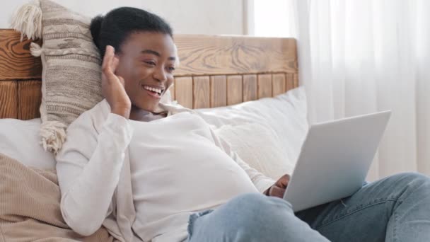 Afrikanische glücklich junge schwangere Frau Freiberuflerin arbeitet einsam zukünftige Mutter Afro-Mädchen mit Laptop macht Videotelefonie Online-Chat-Konferenz von zu Hause aus auf dem Bett liegend winkt hallo spricht Webcam — Stockvideo