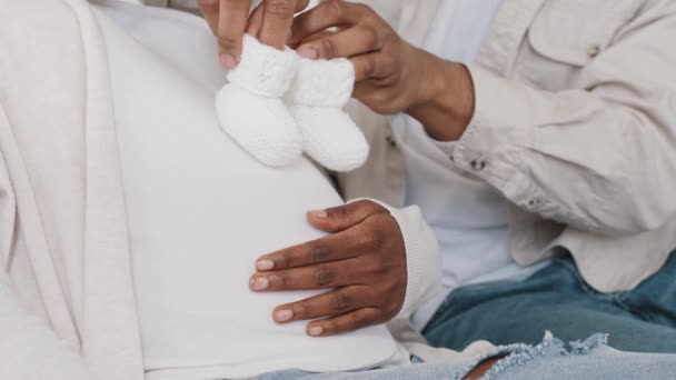 Esperando padres padre irreconocible cuidado marido africano jugar con el vientre femenino embarazada pisando con dulces pequeños zapatos de bebé blancos en la barriga grande de la amada esposa afro negro de cerca, el embarazo — Vídeos de Stock