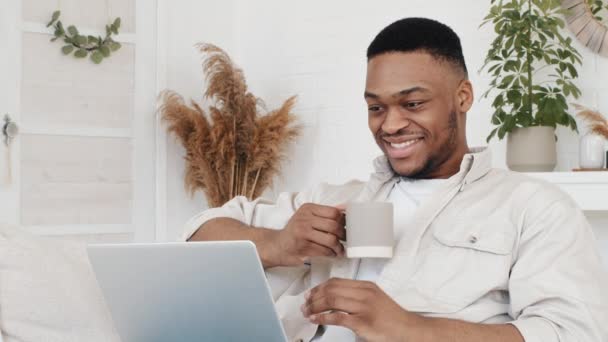 Jovem africano homem freelancer afro estudante preto macho sentado em casa sofá usa aplicativo de computador portátil para estudos de trabalho assistindo filmes lendo notícias navegação rede beber chá café da xícara — Vídeo de Stock