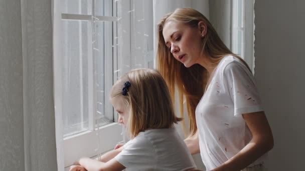 Caucasian young mother babysitter woman caring mom with little daughter child girl looking out window waiting contemplate enjoying sunset isolation in quarantine, small kid points with finger outside — Stock Video