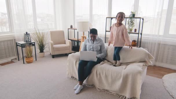 Afro küçük kız çocuğu bebek gürültücü çocuk kanepeye atlıyor kötü davranışlar sergiliyor anne siyah kadın serbest bilgisayar kullanıyor annelik ve karantina sırasında uzaktan kumanda kullanıyor — Stok video