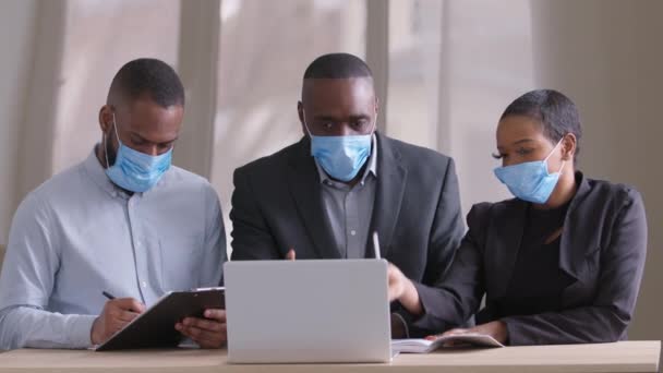 Diversos afro preto equipe de negócios em máscaras protetoras faciais médicas que trabalham no escritório moderno discutindo ideias olhar laptop. Diferentes idades colegas africanos vestindo respiradores conversando sentado à mesa — Vídeo de Stock