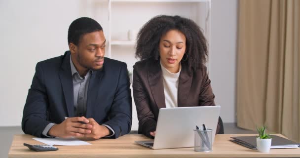 Dwie osoby w biurze licząc wydatki za pomocą kalkulatora afro amerykańska kobieta finansista wprowadzenie danych liczbowych do usługi płatności online aplikacji w laptopie etniczny biznesmen pisze raport na papierze — Wideo stockowe