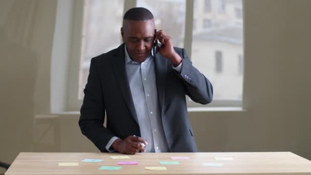 Зрелый бизнес среднего возраста мужчина старший африканский босс afro лидер носит официальный костюм говорить по мобильному телефону с партнерами в чате беспроводной гаджет пишет липкие заметки идеи на бумаге стоя в офисе — стоковое видео