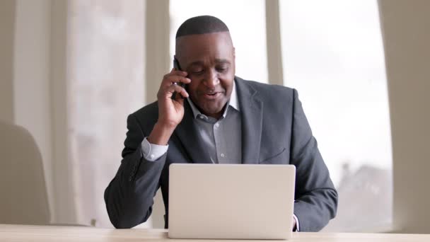 オフィスのノートパソコンで電話に出るアフリカの成熟したビジネスマンに焦点を当てました。職場でクライアントとスマートフォンを話すアフロシニア男性。無線装置携帯電話と呼ばれる黒人男性労働者 — ストック動画