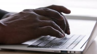 Yakın plan siyah erkek elleri ofis masasında dizüstü bilgisayarda klavye kullanıyor. Tanımlanamayan yetişkin Afrikalı adam içeride bilgisayarla çalışıyor. Bilinmeyen afro işadamı internette geziniyor, kırpılmış kenar görünüm