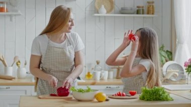 Yetişkin anne ev hanımı kırmızı biberli sebze salatası keser kız çocuk yemek pişirmede annene yardım eder göz makyajı yapmak yerine iki domates tutar anne kadın mutfakta eğlenir