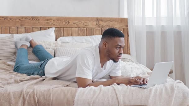 Jovem e bonito homem africano freelancer trabalhando online deitado na cama usa laptop. Focado afro masculino negro cara etnia estudante estudo remotamente a partir aconchegante casa quarto navegação conversando digitação no computador — Vídeo de Stock