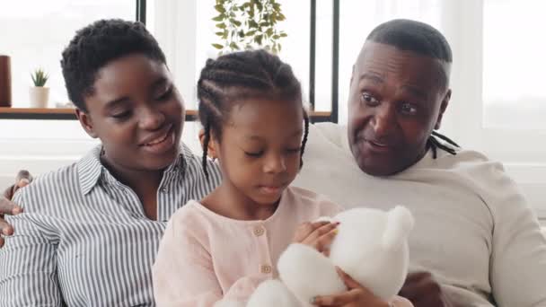 Boldog afrikai család ül a kanapén, anya lánya és nagyapja ölelés csevegés otthon, érett férfi apa megmutatja copfos fonatok kisgyermek fekete gyerek lány, beszél hasonlóság frizurák — Stock videók