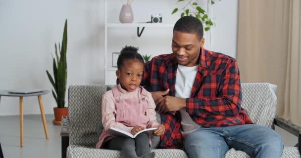 Afro-americano padre single e bambina prescolare seduti insieme sul divano in salotto a fare i compiti, il giovane padre amorevole aiuta la sua adorata figlia con lezioni insegna al bambino a leggere il libro — Video Stock