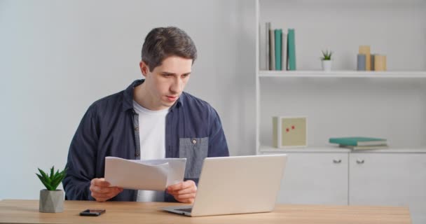 Concentrated caucasian millennial guy businessman manager entrepreneur adult man working sitting at home office looking at laptop reads report checks paper documents enters data into online service — Stock Video
