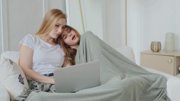 Adult mother with teenager daughter sitting on couch using laptop online browsing network watching evening movie, schoolgirl child girl yawns falling asleep fatigue cuddling mom covered with blanket — Stock Video