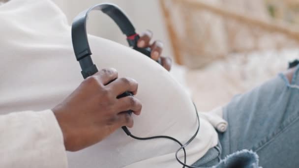 Close-up hovedtelefoner på kvindelige gravide mave. Ugenkendelig afrikansk kvinde vordende mor fremtid afro mor sætter øretelefoner på maven, ufødte barn lytter til musik i livmoderen. Graviditet barsel koncept – Stock-video