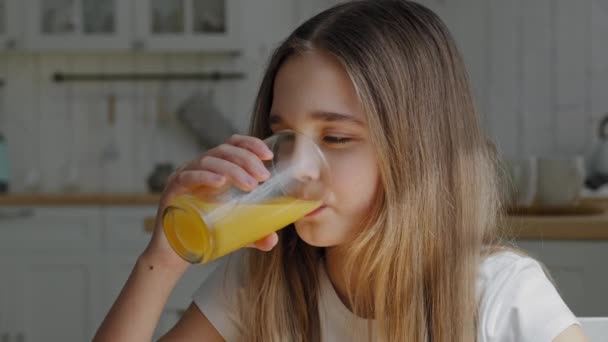 Portrait caucasian little school girl child daughter preteen kid feels thirsty drinks orange juice with tropical fruits vitamins enjoying refreshing taste sitting in kitchen smiling looking at camera — Stock Video