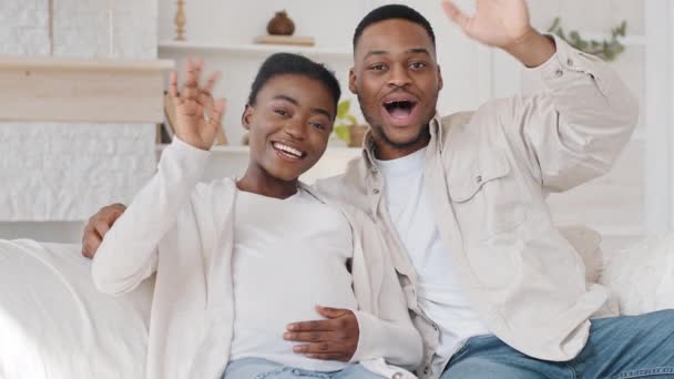 Afro family portrait african pregnant couple cuddling sitting on sofa at home waving hands hello greetings looking at camera talking holding arms on belly sharing news about pregnancy in video call — Stock Video