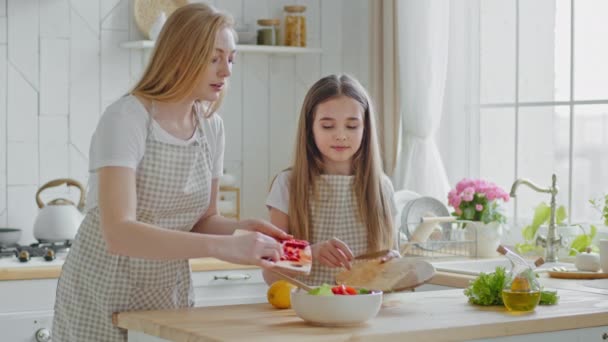 Mother teaches teenage daughter cook, mom with little girl child add red pepper ingredients vegetables into plate with salad baby mix with wooden spoon woman cuts lemon, family cooking in kitchen — Stock Video
