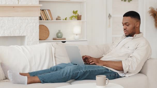 Zaměřený africký student ležící na gauči s notebookem, který si prohlíží webovou studii vzdáleně od domova v karanténě. Afro černošky muž na volné noze pracuje s počítačem sedí na pohovce nakupování on-line — Stock video