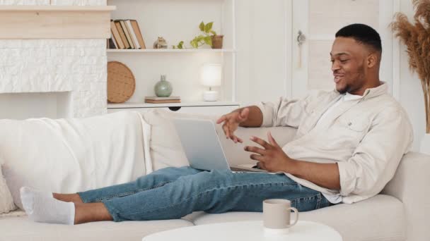 O cara feliz africano está no sofá usando laptop faz conferência de bate-papo de chamada de vídeo online acenando olá para webcam fala remotamente com amigos durante a quarentena. Afro homem vídeo a partir de casa em lockdown — Vídeo de Stock