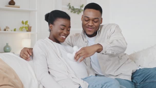 Afroamerykańska para rodzina przyszłość oczekując rodziców siedzących na kanapie afro czarny mąż mężczyzna głaskanie żona ciężarnych brzuch z cute małe dziecko białe buty przytulając ukochaną kobietę — Wideo stockowe