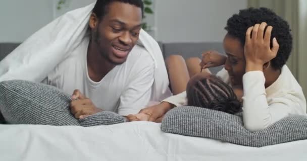 Afrikanische Familie liegt auf Sofa auf grauen Kissen, bedeckt mit weißer warmer Decke Afro-Mutter und ethnische Mädchen Tochter Kind im Gespräch mit glücklichen schwarzen Vater hält Smartphone-Handy — Stockvideo