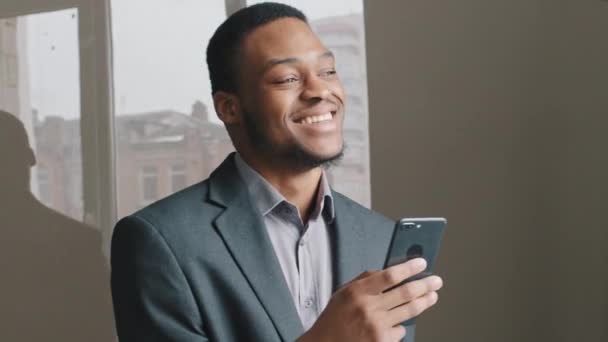 Emocionado hombre negro que trabaja en la aplicación digital mantenga teléfono inteligente leer buenas noticias de Internet, chico africano feliz mirar la pantalla del teléfono encantado por el éxito de la victoria. Millennial empresario usuario de tecnología móvil — Vídeo de stock