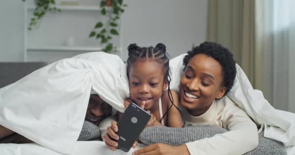 Funny Afro american glad etnisk sort familie liggende på sofaen dækket med tæppe, lille afrikaner datter pige tager fotooptagelse video på mobiltelefon kamera, mor poserende, far gemmer griner – Stock-video