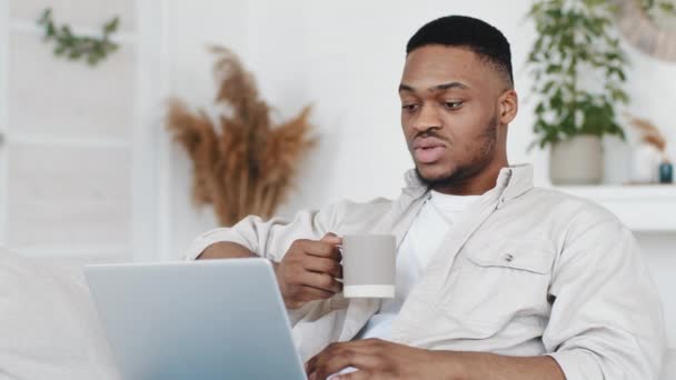 Homem africano jovem afro estudante étnico preto macho sentado em casa segurando xícara com chá quente café olhando para tela do laptop lê em voz alta fala por vídeo bate-papo aprende palavras texto, quarentena e-learning — Vídeo de Stock